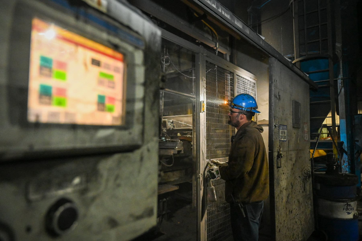 A factory worker wearing a blue hard hat and gloves operates machinery in an industrial setting with control panels and equipment around.